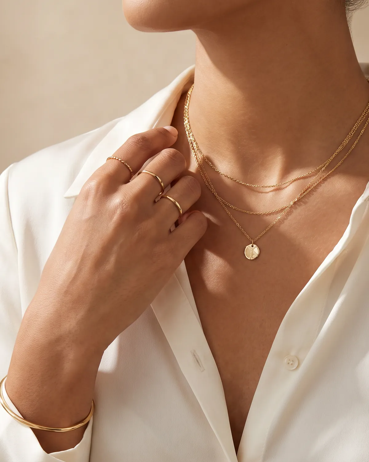 A hand resting at the collar of a cream silk shirt, wearing stacked gold rings, a bangle and layered gold necklaces