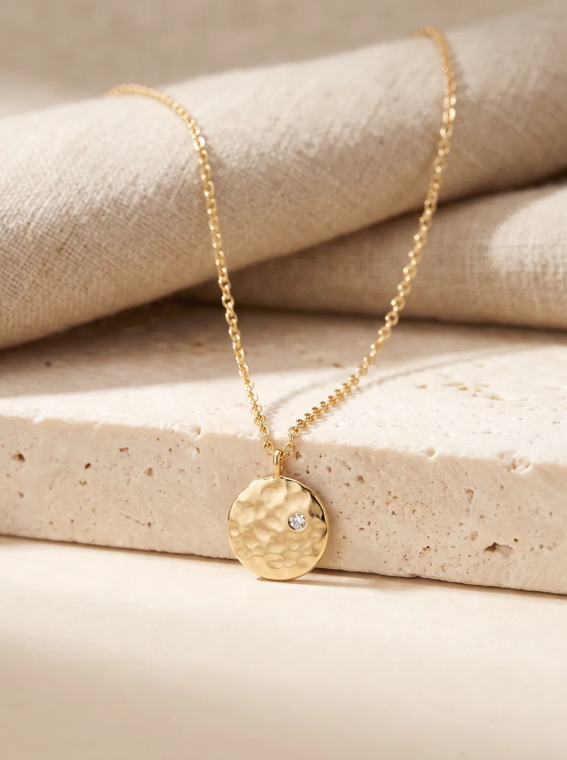 The Solstice pendant, a small hammered gold disc set with one diamond, on a fine chain draped over travertine and linen